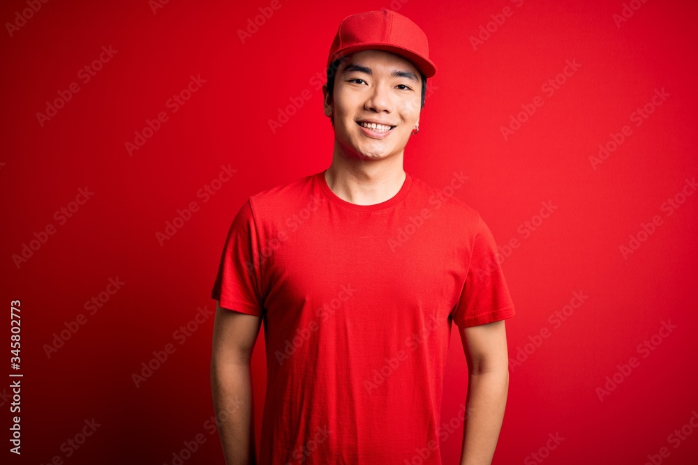 Young handsome chinese delivery man wearing cap standing over isolated red background with a happy and cool smile on face. Lucky person.