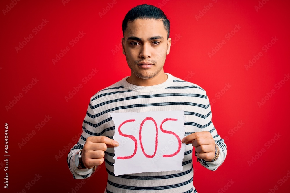 Young brazilian man with problem holding banner with sos message over red background with a confident expression on smart face thinking serious Young brazilian man with problem holding banner with sos message over red background with a confident expression on smart face thinking serious