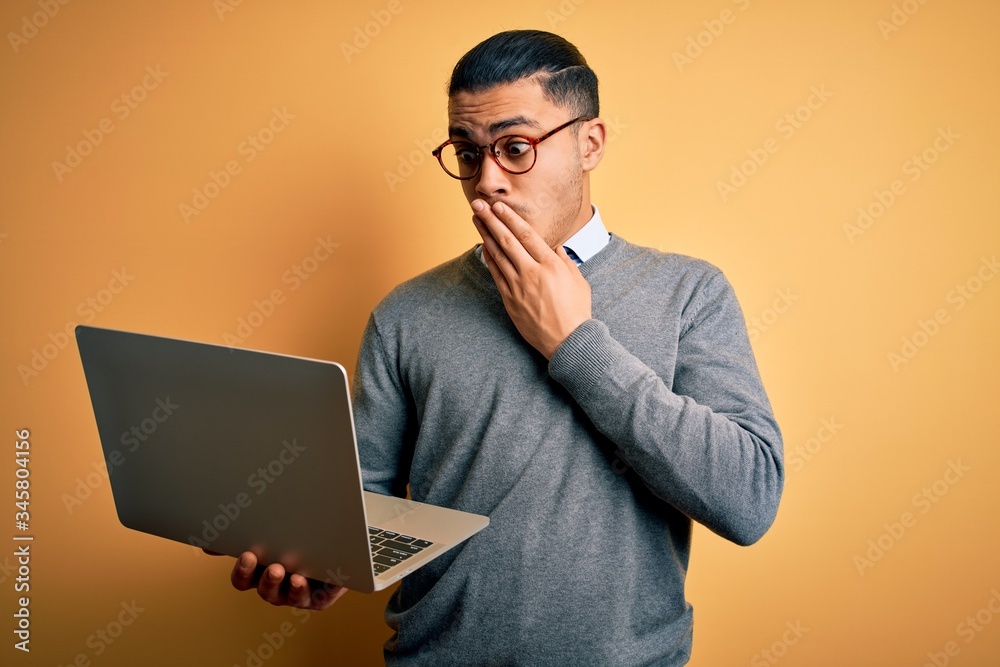 Young brazilian businessman working using laptop standing over isoltated yellow background cover mouth with hand shocked with shame for mistake, expression of fear, scared in silence, secret concept