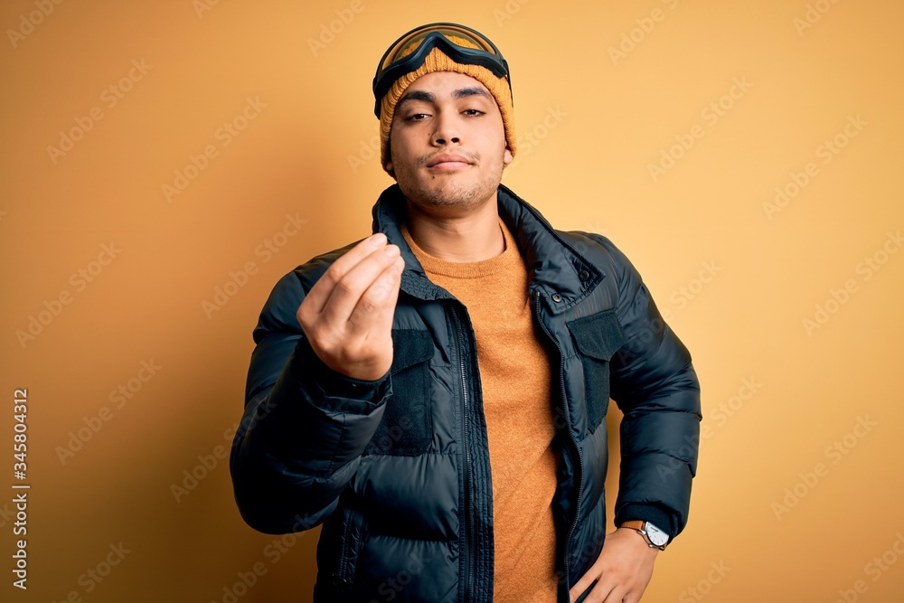Young brazilian skier man wearing snow sportswear and ski goggles over yellow background Doing Italian gesture with hand and fingers confident expression Young brazilian skier man wearing snow sportswear and ski goggles over yellow background Doing Italian gesture with hand and fingers confident expression