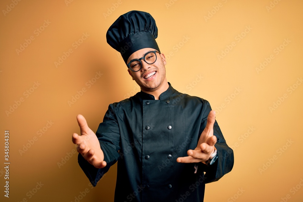 Young brazilian chef man wearing cooker uniform and hat over isolated yellow background smiling cheerful offering hands giving assistance and acceptance. Young brazilian chef man wearing cooker uniform and hat over isolated yellow background smiling cheerful offering hands giving assistance and acceptance.