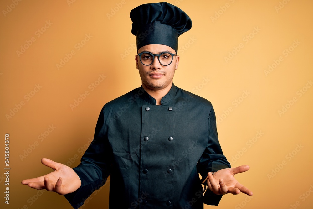 Young brazilian chef man wearing cooker uniform and hat over isolated yellow background clueless and confused with open arms, no idea concept. Young brazilian chef man wearing cooker uniform and hat over isolated yellow background clueless and confused with open arms, no idea concept.