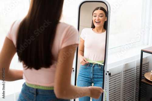 Happy Girl Measuring Waist Standing In Front Of Mirror Indoors