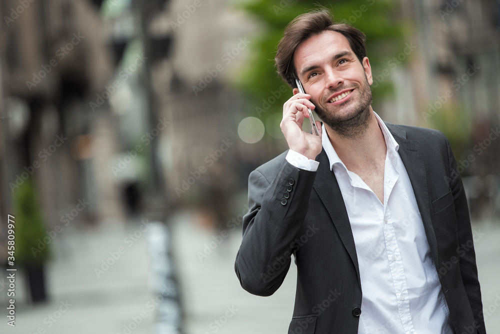 Attractive young businessman having a pleasant phone talk while walking in the urban part of the city Attractive young businessman having a pleasant phone talk while walking in the urban part of the city