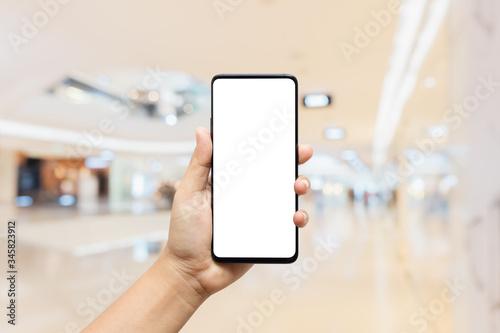Mockup, person hand holding blank white screen mobile smart phone with blurred background of modern luxury department store for your advertisement artwork