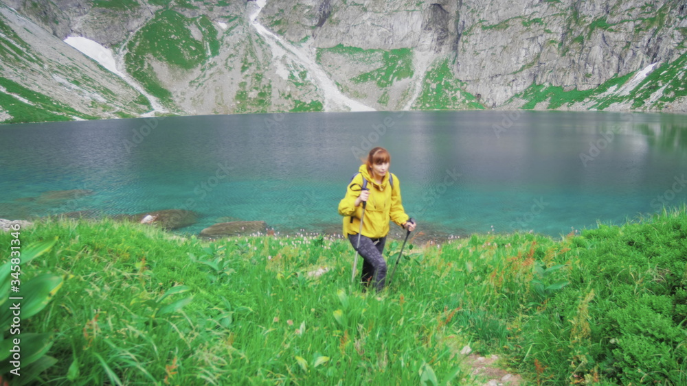 A young tourist woman walks upstairs with nordic walking uphill from a beautiful mountain lake and enjoys her adventure. Sports recreation activity. Summer vacation tourism. Slow motion 4K video. A young tourist woman walks upstairs with nordic walking uphill from a beautiful mountain lake and enjoys her adventure. Sports recreation activity. Summer vacation tourism. Slow motion 4K video.