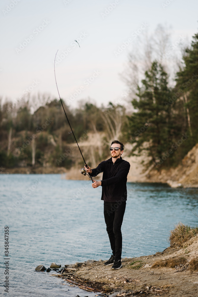 Fisherman with rod throws bait into the water on river bank. Fishing for pike, perch, carp. Man catching fish, fishing on lake, pond with text space. Wild nature. The concept of rural getaway. Fisherman with rod throws bait into the water on river bank. Fishing for pike, perch, carp. Man catching fish, fishing on lake, pond with text space. Wild nature. The concept of rural getaway.