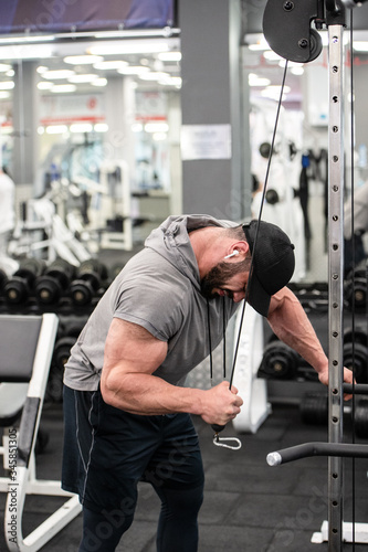 strong young bearded male in cap and sleeveless hoodie heavy weight training in sport gym using exercise fitness equipment listening music on wireless headphones © ruslanshug