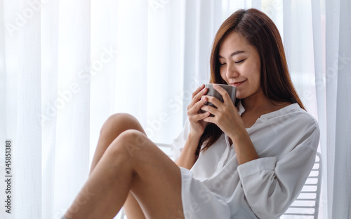 A beautiful asian woman drinking hot coffee in a white bedroom at home in the morning
