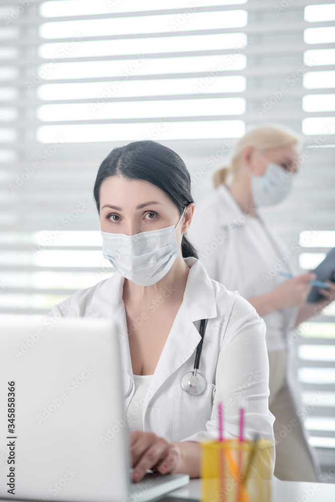 Masked woman with stethoscope behind laptop, blonde at window. Masked woman with stethoscope behind laptop, blonde at window.