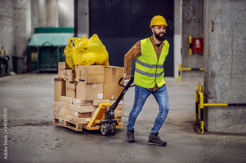 Full length of smiling hardworking bearded worker in vest, with safety helmet on head pulling a pallet truck with boxes, sack and building material. Building in construction process interior.