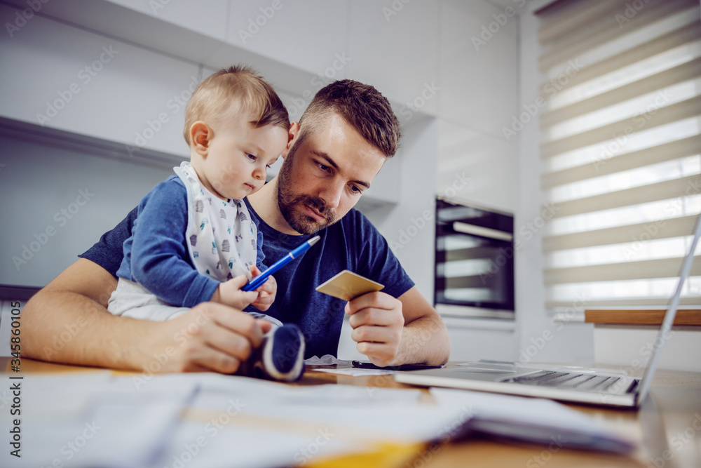 Young bearded pensive bearded young father holding credit card and preparing to pay bills by laptop. His little helper sitting on table. Young bearded pensive bearded young father holding credit card and preparing to pay bills by laptop. His little helper sitting on table.