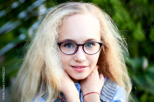 Pretty teenage girl 14-16 year old with curly long blonde hair and in glasses in the green park in a summer day outdoors. Beautiful portrait
