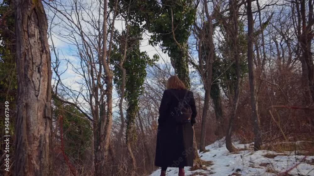 A blond girl with black clothes walking in between trees in a forest in the snow during wintertime