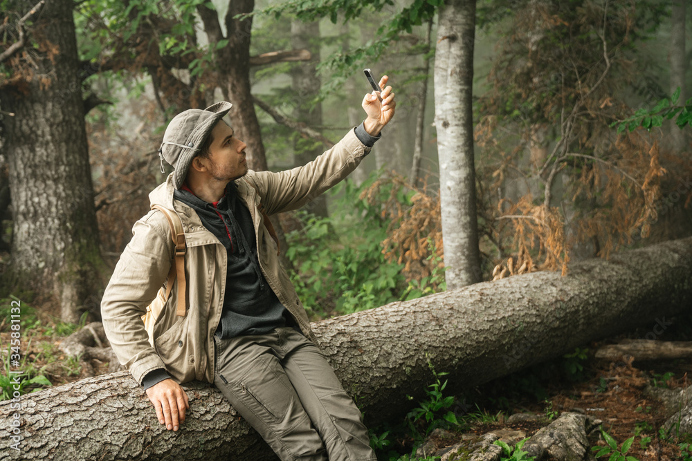 man in hiking clothes holds a smartphone in his hand while sitting on a log in the forest and taking a selfie photo or using mobile communication to chat online. Man and navigator.