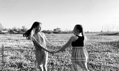 two teenage sisters in summer dresses holding hands and spinning in dandelion meadow