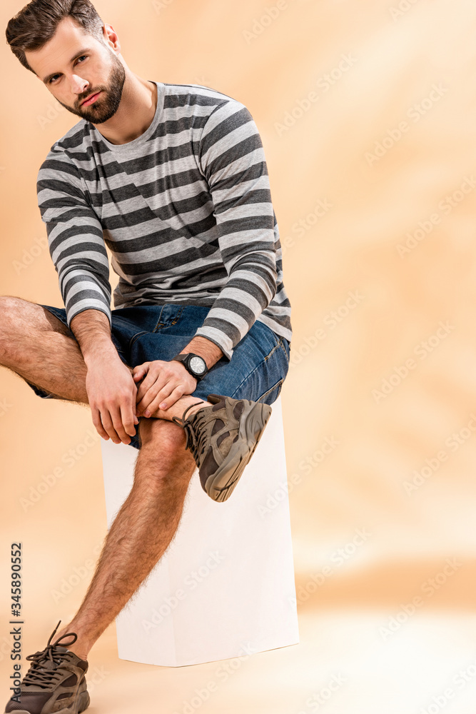 handsome bearded man in striped sweatshirt sitting on white cube on beige