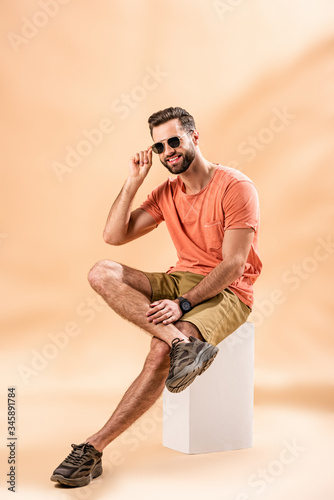 smiling handsome young man in shorts, summer t-shirt and sunglasses sitting on white cube on beige photo