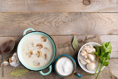 Turquoise ceramic pan with mushroom cream soup, cup with sour cream, bowl with fresh champignons, spoon and spesies on burlap on wooden background. photo