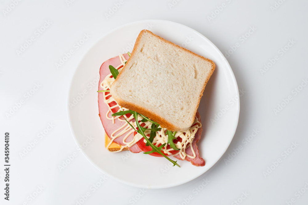 Tasty sandwich with ham or balyk, tomato, arugula, cheese, mayonnaise and white bread for toast on a plate on a white background view from the top flat lay, copy space
