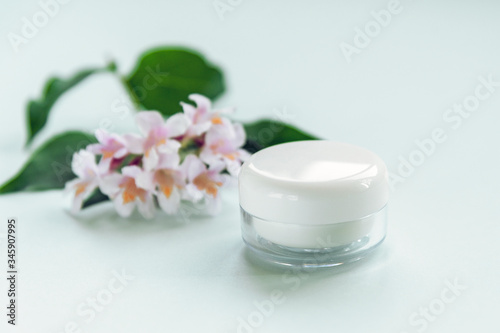 Cosmetic cream in white plastic jar on the grey background with flowers. Unbranded package