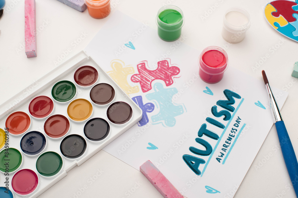 top view of card with Autism Awareness Day lettering and painting of puzzle on white with paints, paint brush and chalks top view of card with Autism Awareness Day lettering and painting of puzzle on white with paints, paint brush and chalks