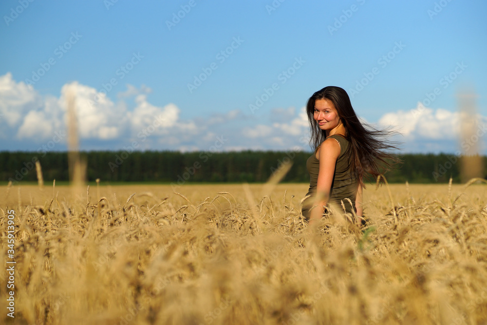 Beautiful woman with long hear standing waist-deep in wheat field, turning head backwards. Hands touching beige ears Beautiful woman with long hear standing waist-deep in wheat field, turning head backwards. Hands touching beige ears
