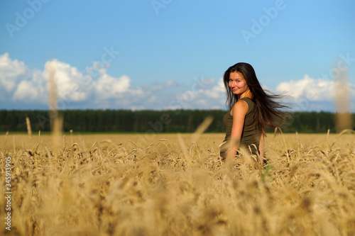 Beautiful woman with long hear standing waist-deep in wheat field, turning head backwards. Hands touching beige ears