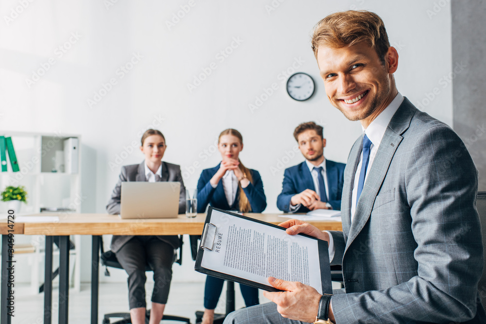 Selective focus of smiling employee with resume looking at camera near recruiters in office Selective focus of smiling employee with resume looking at camera near recruiters in office