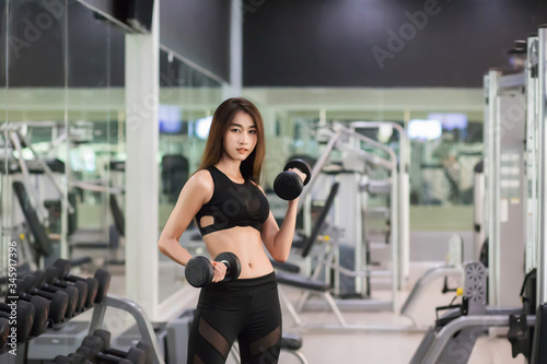 A beautiful Asian woman, Thai, is wearing a sexy black workout outfit. She is lifting a dumbbell in a gym full of exercise equipment.