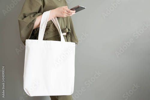 Blank white tote bag canvas fabric with handle mock up design. Close up of woman holding eco or reusable shopping bag and using smartphone near grey wall. No plastic bag and ecology concept. photo