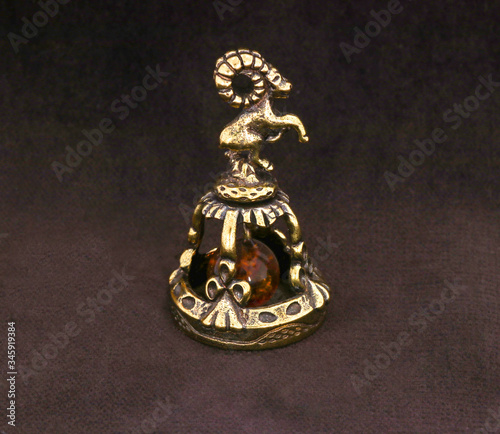 Amber souvenir. A miniature metal bell in the form of a zodiac sign "Capricorn" with an amber element. Background is dark brown or white (isolated image).