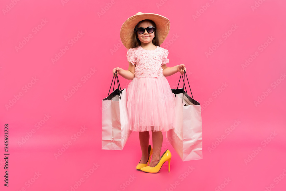 Happy lovely moments of shopping time with cute little girl in dress standing in mother`s big shoes with white packages in hands isolated on pink background