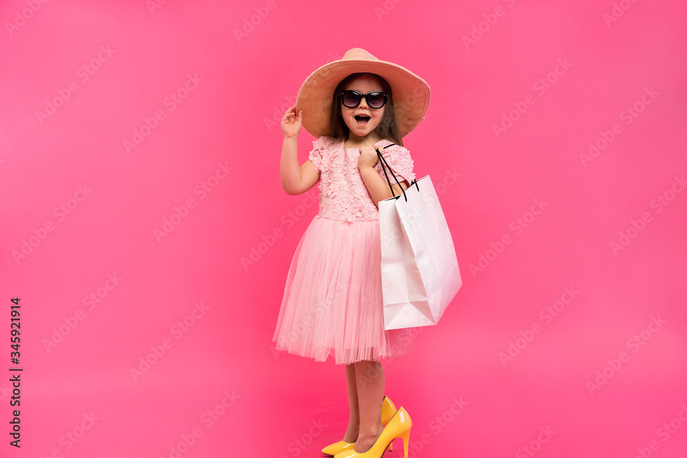 Happy lovely moments of shopping time with cute little girl in dress standing in mother`s big shoes with white packages in hands isolated on pink background