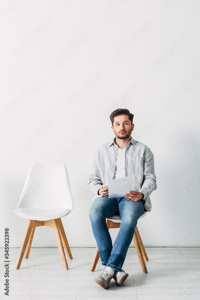 Employee with resume and smartphone looking at camera on chair in office Employee with resume and smartphone looking at camera on chair in office
