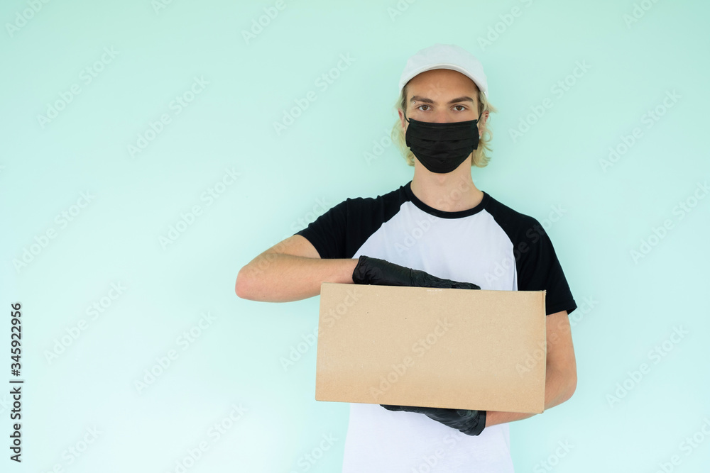 a guy in a white T-shirt with a black short sleeve and a white cap, in a black protective mask holds a cardboard box in his hands. Home delivery during quarantine