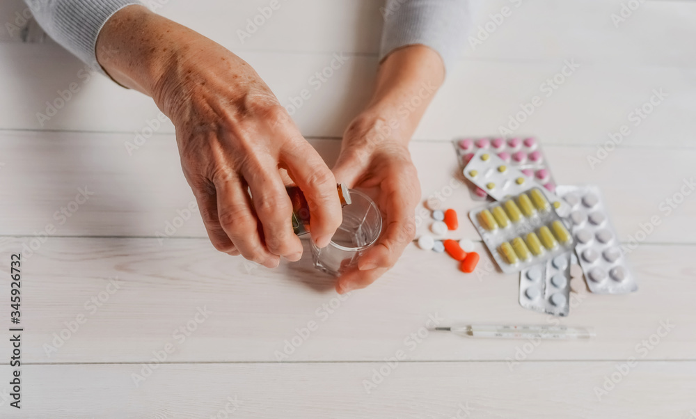 Obraz premium Senior hands with medicine bottle putting drops in a glass, pills, drugs, thermometer on a table. Woman wrinkled hands, colorful tablets, wooden background. Health care for the elderly, old age.