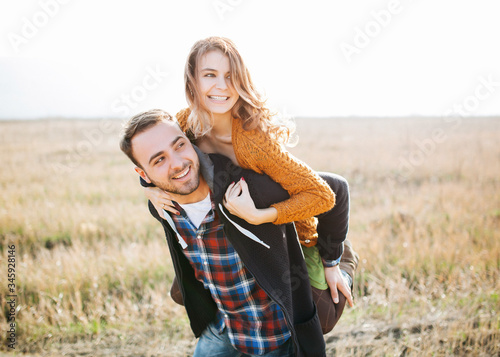 Young cheerful couple of man and woman playing in an open field, piggy-back ride, backlit. Shallow depth of field.