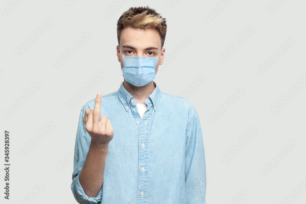 Portrait of anger young man with surgical medical mask in light blue shirt standing with middle finger fuck sign and looking at camera with serious or poker face. indoor studio shot on grey background Portrait of anger young man with surgical medical mask in light blue shirt standing with middle finger fuck sign and looking at camera with serious or poker face. indoor studio shot on grey background