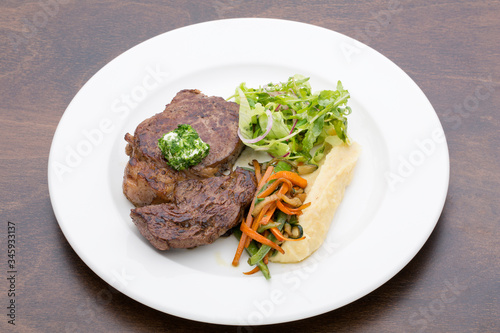 A roasted steak with mashed potatoes, fried vegetables and lettuce in a white ceramic plate on a wooden table