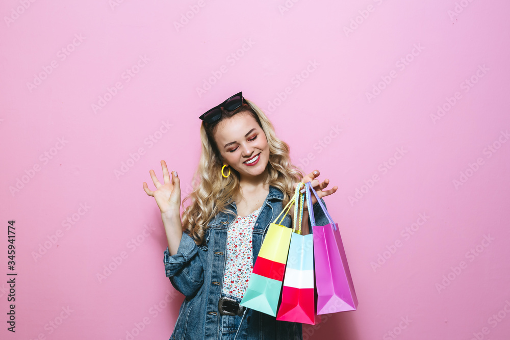 Portrait of a young blond girl holding shopping bags and showing gestures on pink background