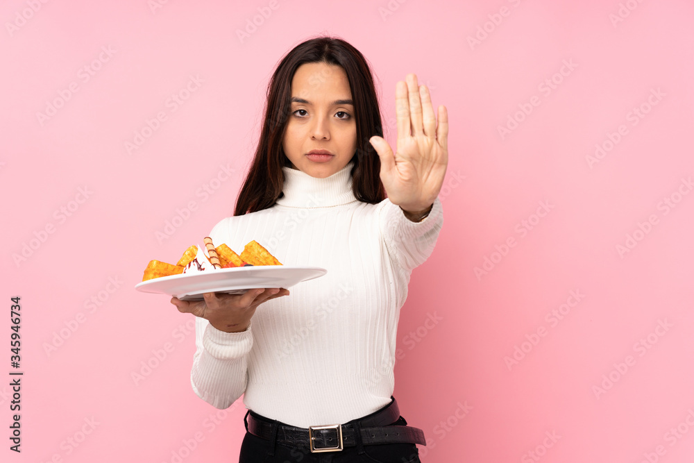 Young brunette woman holding waffles over isolated pink background making stop gesture Young brunette woman holding waffles over isolated pink background making stop gesture