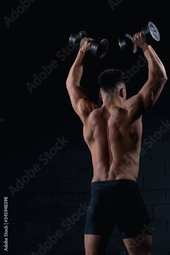 sexy sporty man with his back to the camera holding dumbbells near his head where the delta-shaped trapezoidal three-arm shoulder and widest shoulder muscles stand out, place for text.