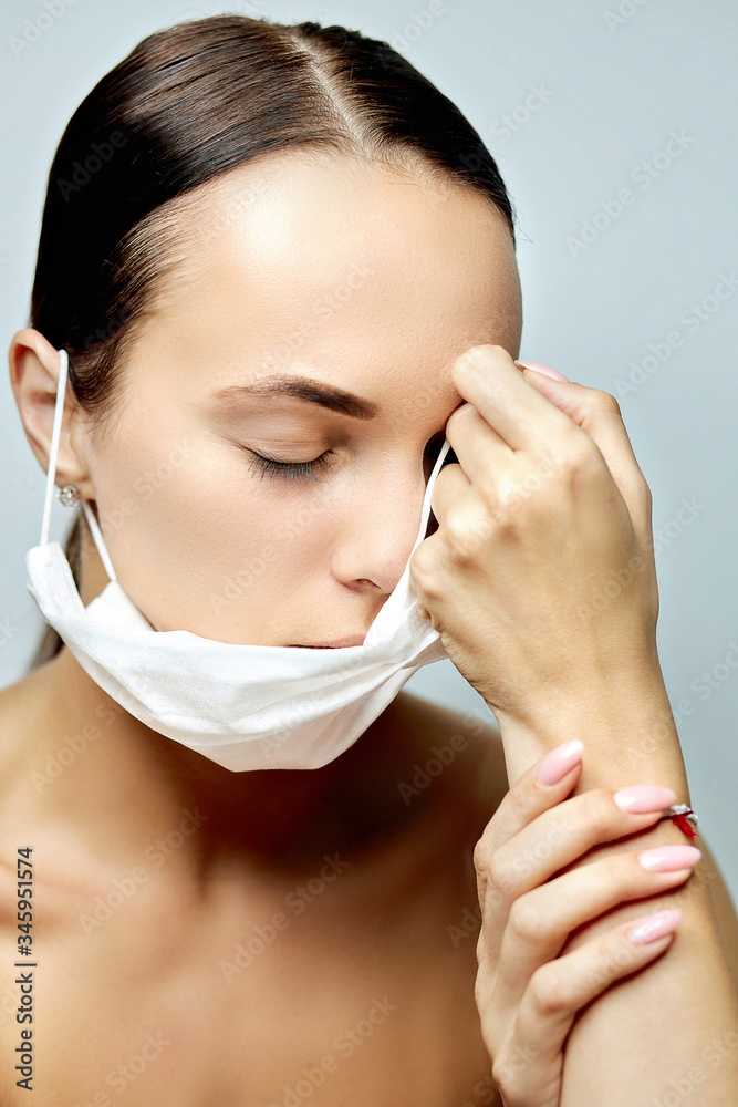 young brunette girl looking down with a tired look with a flat white medical disposable mask on a long neck near the collarbone and well-groomed hands with pink manicure on a gray background