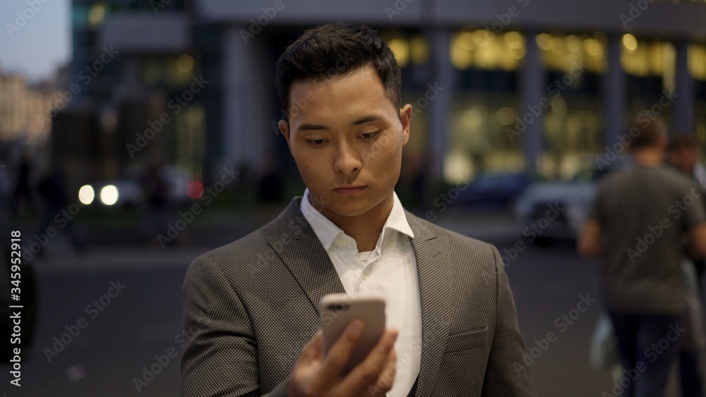 Front view of a young asian businessman, leading correspondence on the phone. Financial business area. Night time at business city. Front view of a young asian businessman, leading correspondence on the phone. Financial business area. Night time at business city.