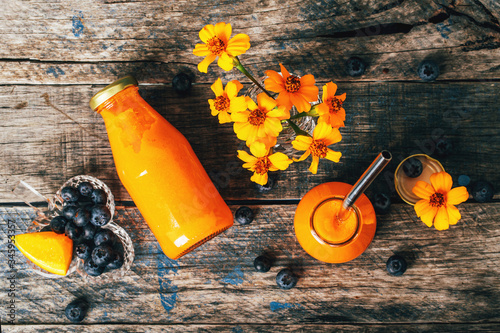 Orange and carrot smoothie in glass bottles with stainless straw and crystal small vases with Chernobrivtsi flowers and blueberries on rustic grunge wooden background. photo