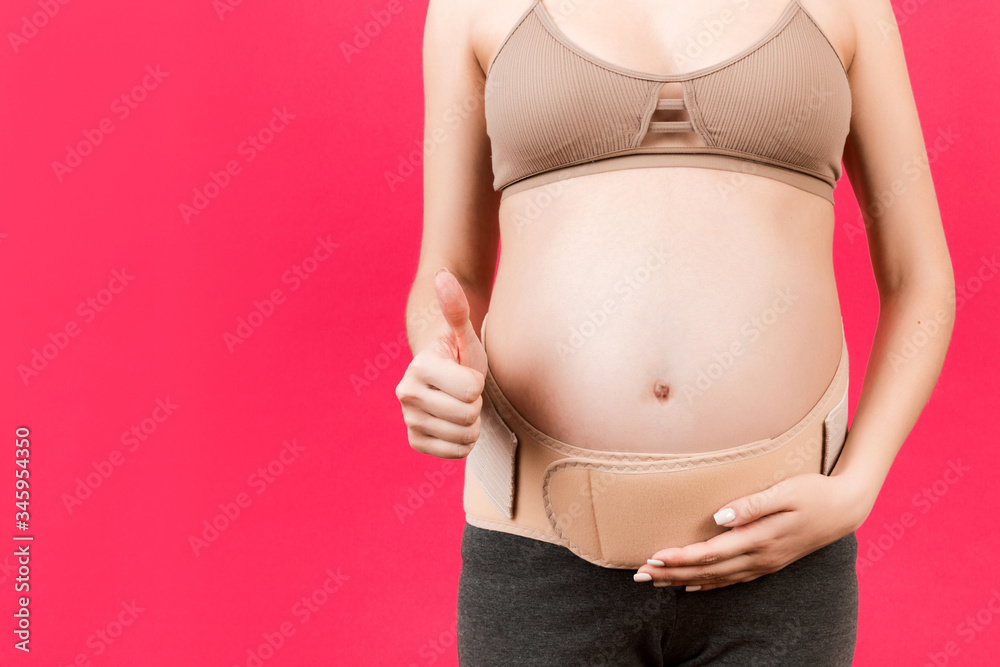 Close up of pregnant woman wearing pregnancy corset against backpain and  showing thumb up gesture at pink background with copy space. Orthopedic  abdominal support belt concept Photos