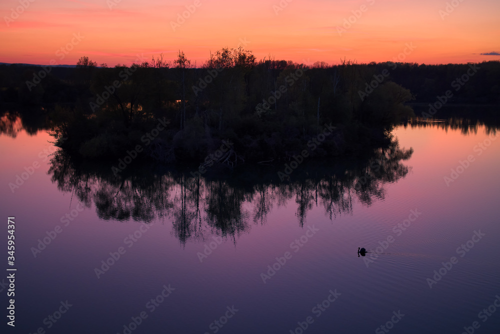 Aerial, after sunset view of silhouette of small isolated island with swan, reflecting in water against colorful orange sunset. Orange and violet colors. Tranquil spring nature.