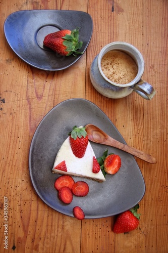 Strawberry cheesecake on dark triangle plate with wooden spoon on wooden table. Chocolate milk with cinnamon and some strawberries on the side. Closeup, vertical, top view, flat lay.
