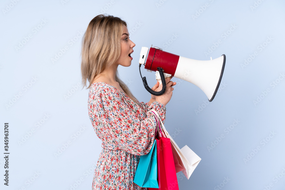 Teenager Russian girl with shopping bag isolated on blue background shouting through a megaphone Teenager Russian girl with shopping bag isolated on blue background shouting through a megaphone